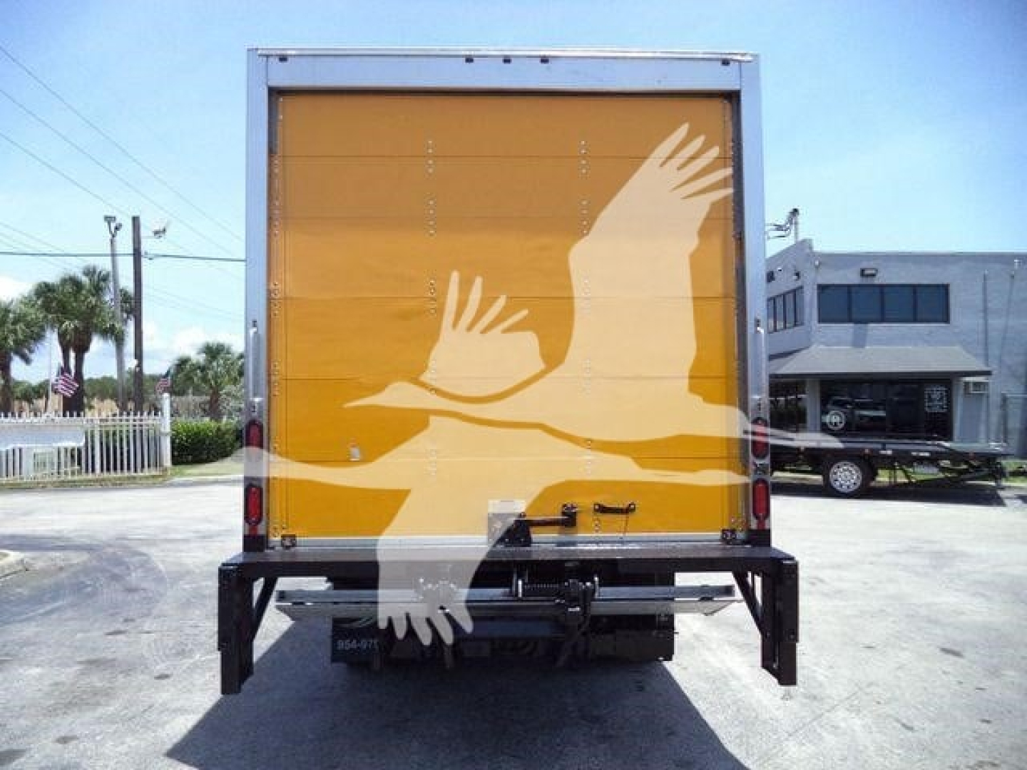 Isuzu NPR HD box truck rear view. Yellow cargo area with white bird graphic. Tail lights on sides, bumper with rear frame visible.