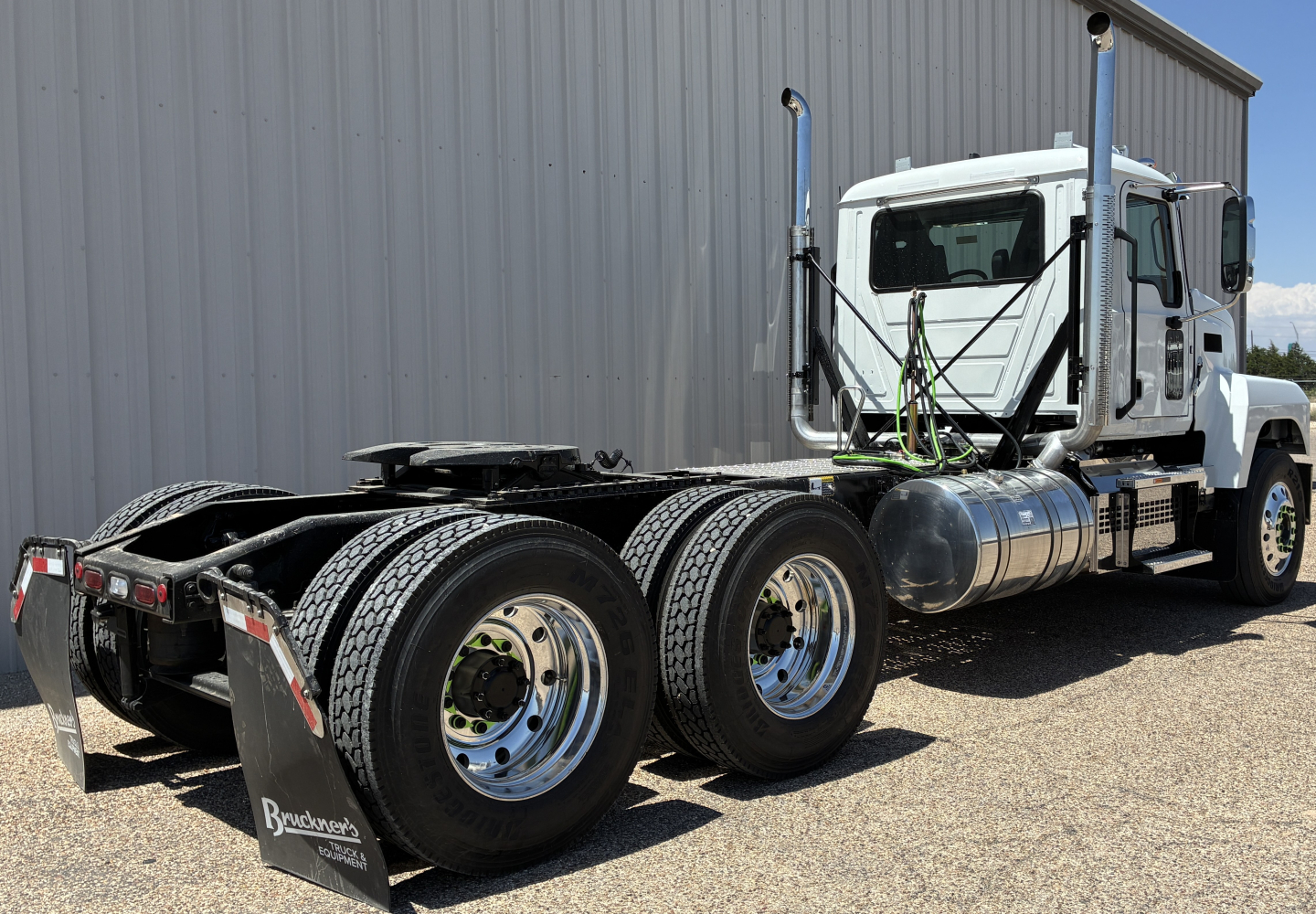 New Mack PI64T conventional daycab tractor showing chassis and dual rear axles, fuel tanks, chromed wheels, and sleeper cab window.