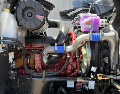 Engine bay of new 2027 Mack PI64T showing coolant reservoir, purple coolant reservoir cap, red engine block, interconnecting hoses, and radiator Fan assembly.