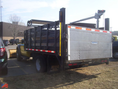 Ford F-550 stake bed truck with plow in rear, metal side rails, and flatbed with tailgate; utility equipment and hitch visible.