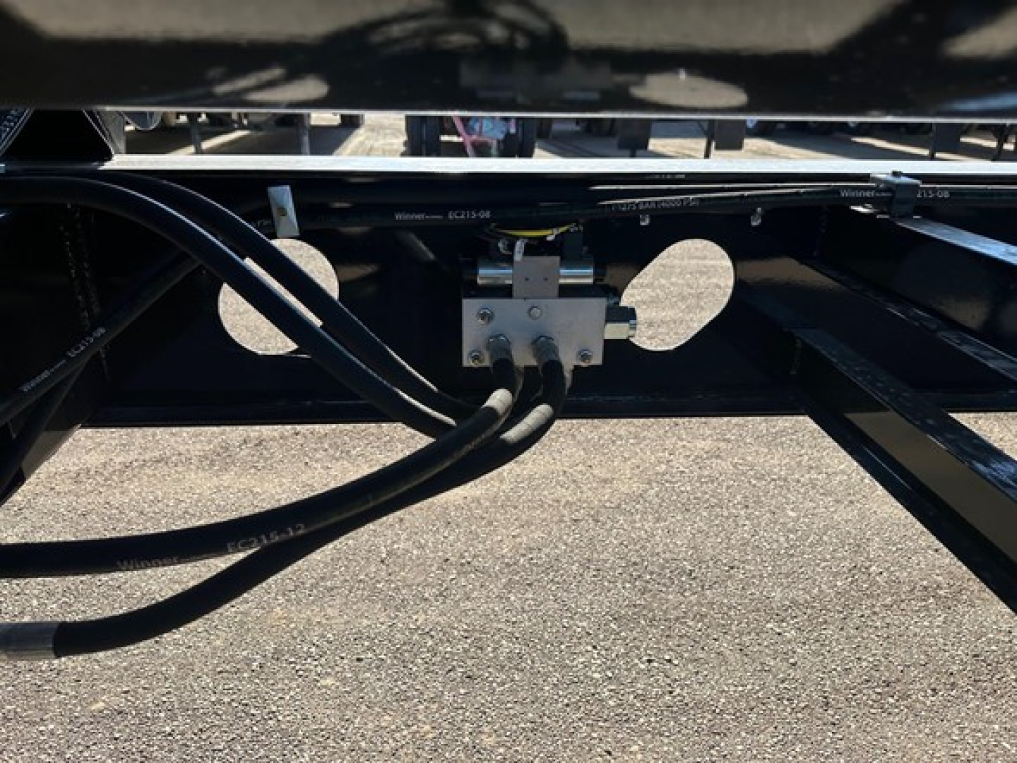 Trailer underside showing hydraulic lines and a rectangular metal valve block with bolts and hoses attached.