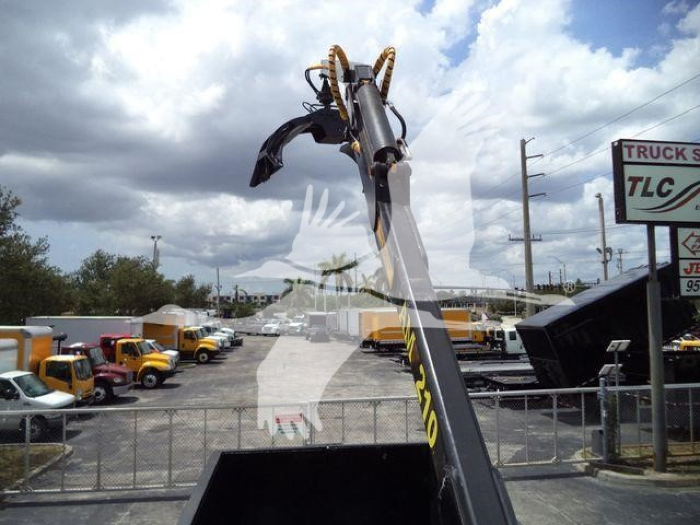 Front portion of a 2019 International 4300 grapple truck showing the crane arm and grapple at the top with a fleet of yellow and white trucks behind, overcast sky.