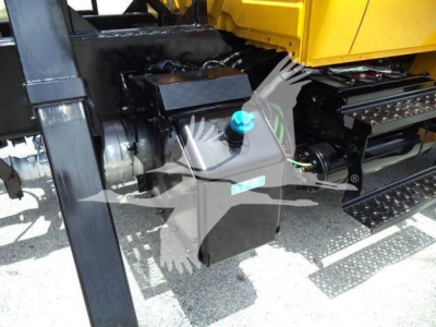 Hydraulic system with black auxiliary tank and blue cap near a yellow grapple truck. Visible hoses, frame, and side steps.