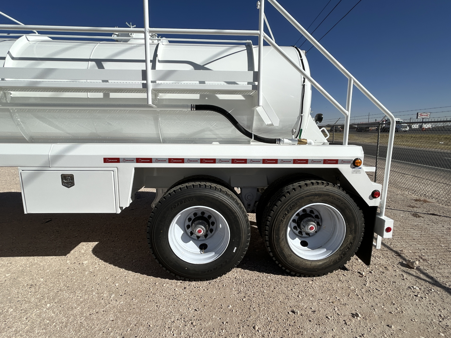Trailer side view showing dual rear wheels, white tanker with railing, storage box, and red reflective strip along the side.