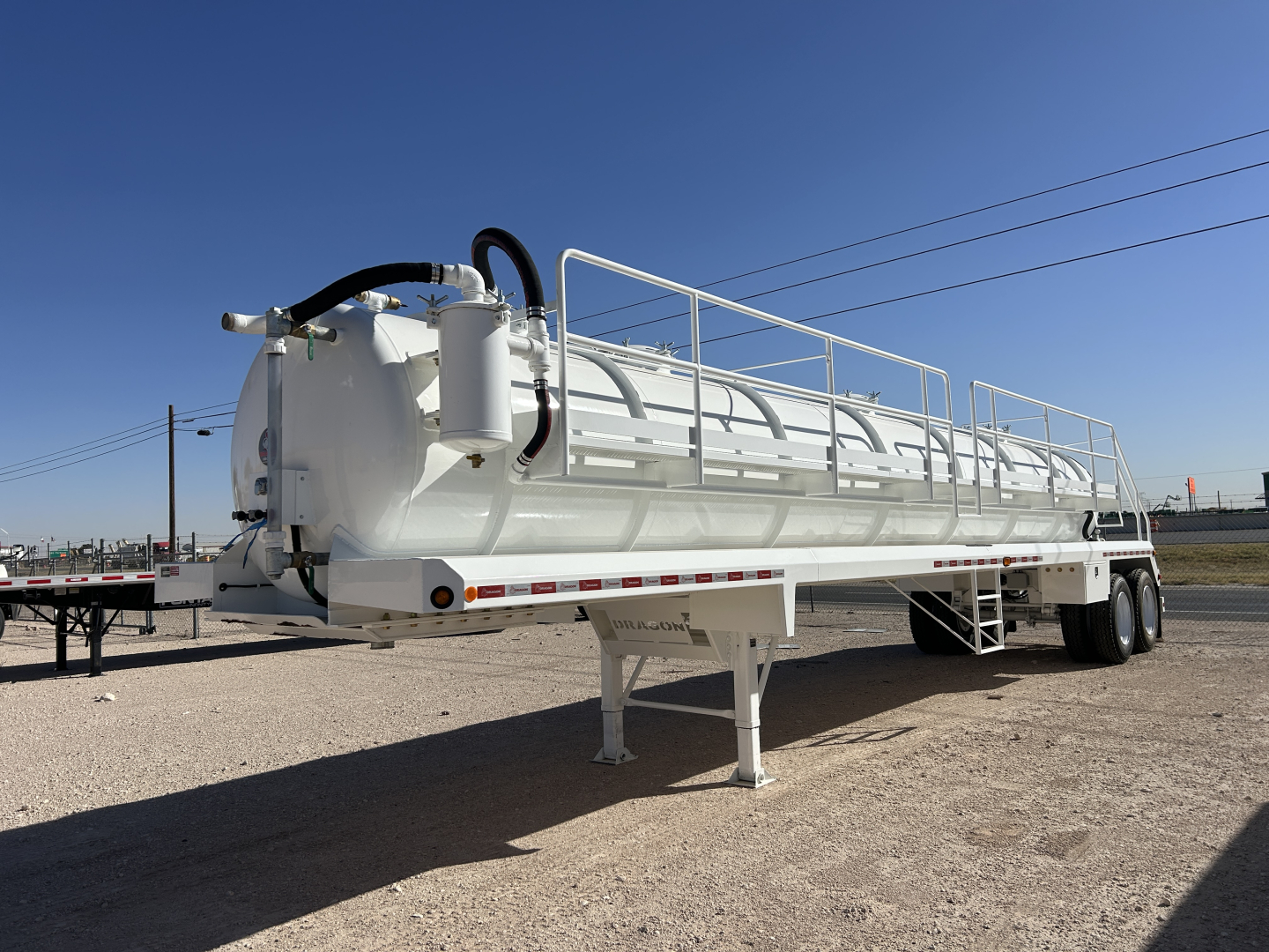 Dragon 130BBL vacuum trailer showing white cylindrical tank with railing, dual-axle wheels, and support legs. Visible hoses and pump attachments at front, reflective strip along bottom edge.