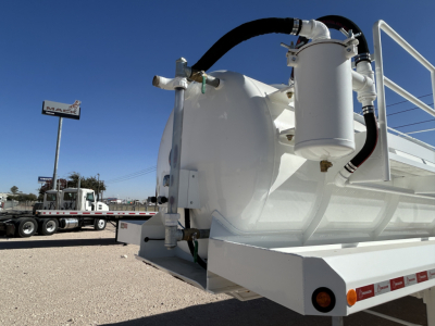 Industrial trailer tank with hoses and fittings; white cylindrical tank mounted on a flatbed trailer, with hoses connected to a vertical filter canister; front-left view showing rear wheel and reflective markers.