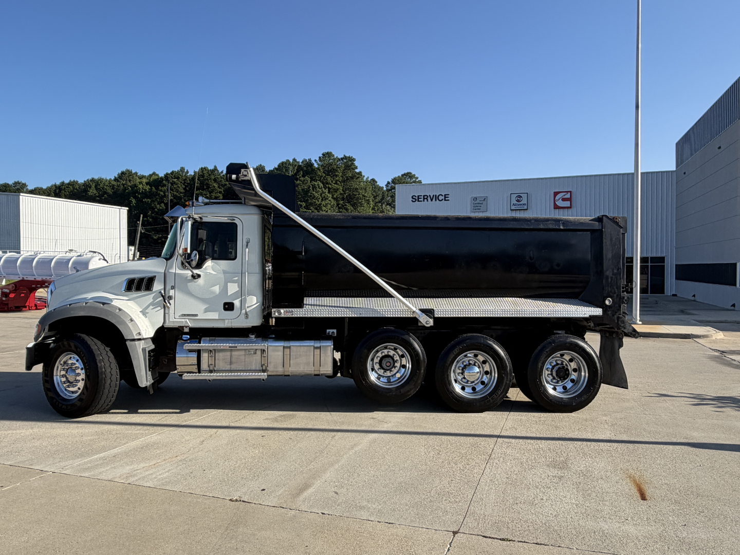 Dump truck 2024 Mack GR64F side view showing large black dump bed and chrome fuel tanks with six wheels and ladder, VIN 1M2GR2GC6RM040889 visible on listing.