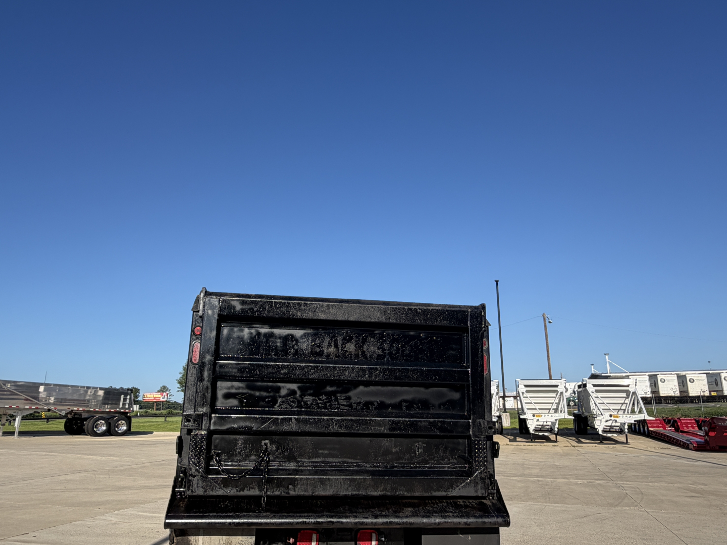Rear view of a black dump truck bed with white lettering or markings in the center, at a dealership lot with other trailers in the background.