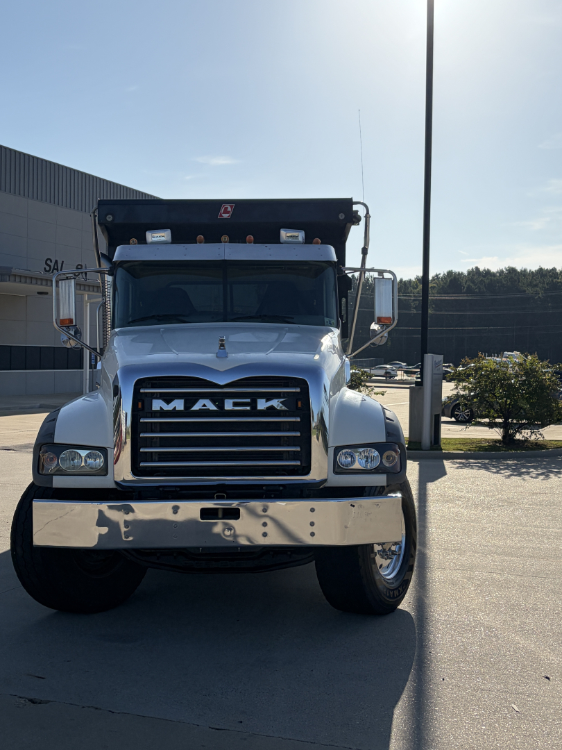 Mack dump truck front view showing large chrome bumper, black grille with MACK letters, dual headlights, and a raised dump bed in a parking lot.