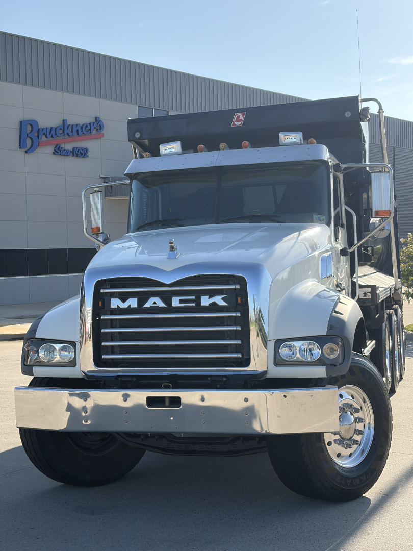 Mack GR64F dump truck front-facing view showing chrome grille with MACK lettering, dual headlights, large bumper, and dump bed in background.