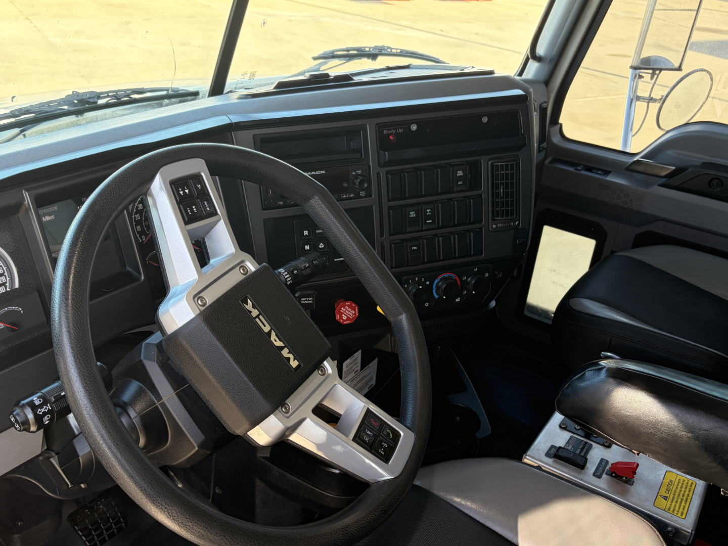 Truck cockpit interior showing steering wheel with Mack logo, dashboard with gauges and controls, center console, driver seat, and pedals.