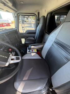 Truck cab interior of a 2024 Mack GR64F showing driver seat, passenger seat, dashboard with controls, steering wheel, and center console; windows reveal exterior yard.