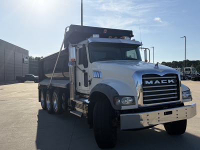 Mack GR64F dump truck in driveway with chrome grille and large front bumper, three rear axles, white cab, and gray dump bed.