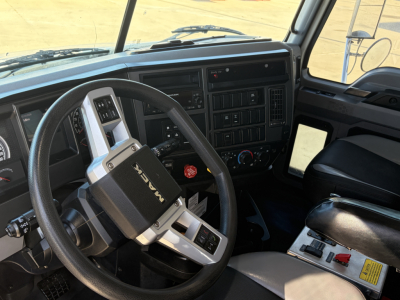 Truck cockpit interior showing steering wheel with Mack logo, dashboard with gauges and controls, center console, driver seat, and pedals.