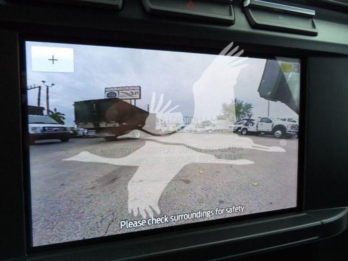 Dashboard screen showing a rear camera view with overlay safety graphics (bird silhouette) and the message 'Please check surroundings for safety.'