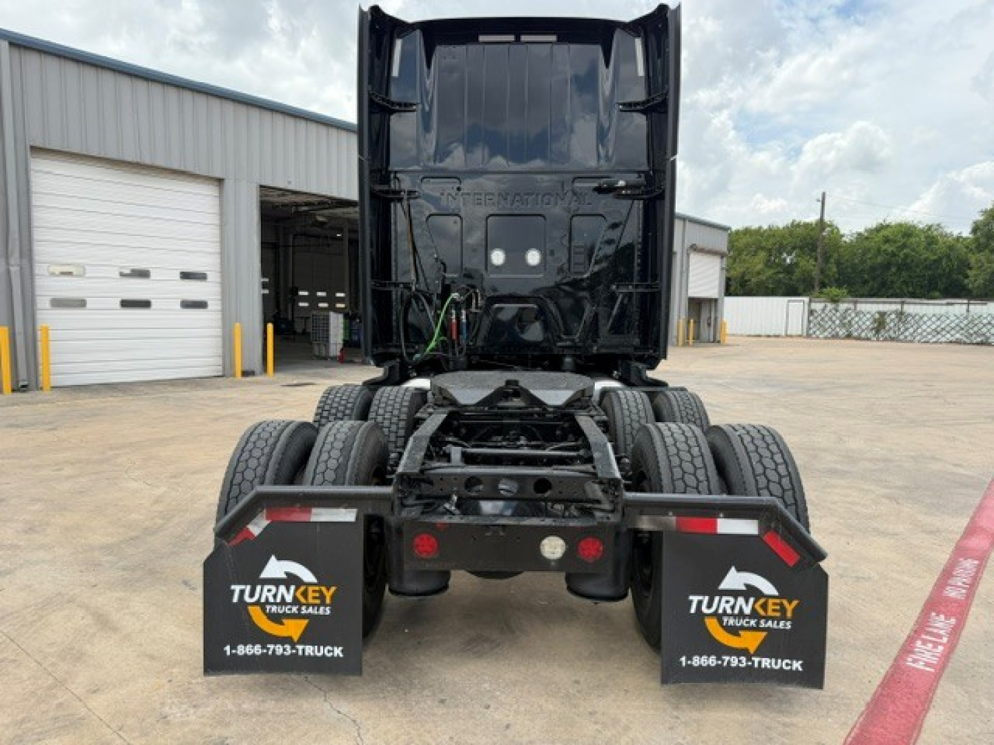 Tractor-trailer cab seen from rear showing fifth-wheel and dual rear axles, black truck frame, Turnkey Truck Sales mud flaps, and dealership lot.