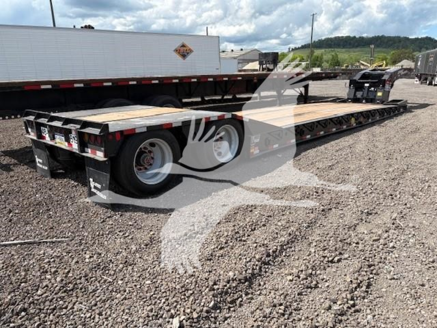 Lowboy trailer showing long flatbed with dual rear axles and white wheels. Visible are wheel wells, trailer deck, and rear bumper lights.