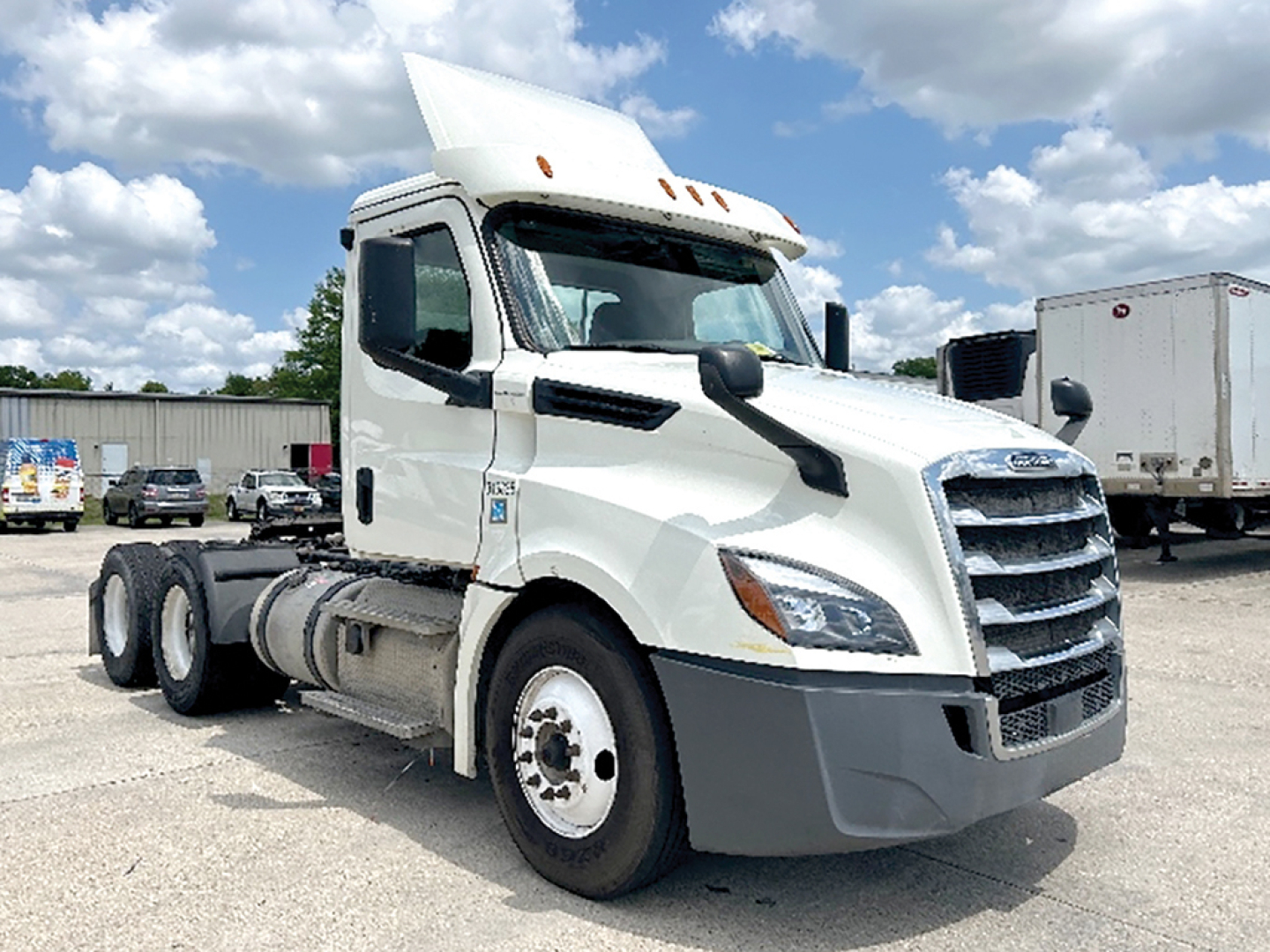Truck cab of a white Freightliner Cascadia126 semi-truck in a dealership lot with blue sky and clouds, 3AKJHLDV7JSJW9739 visible on VIN plate.