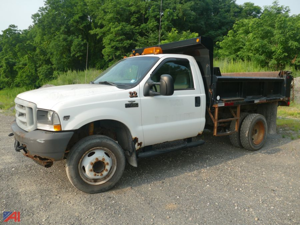 Ford F-450 Super Duty dump truck in service condition, white cab with orange rooftop beacon, black dump bed, gravel lot, visible rust on front bumper.