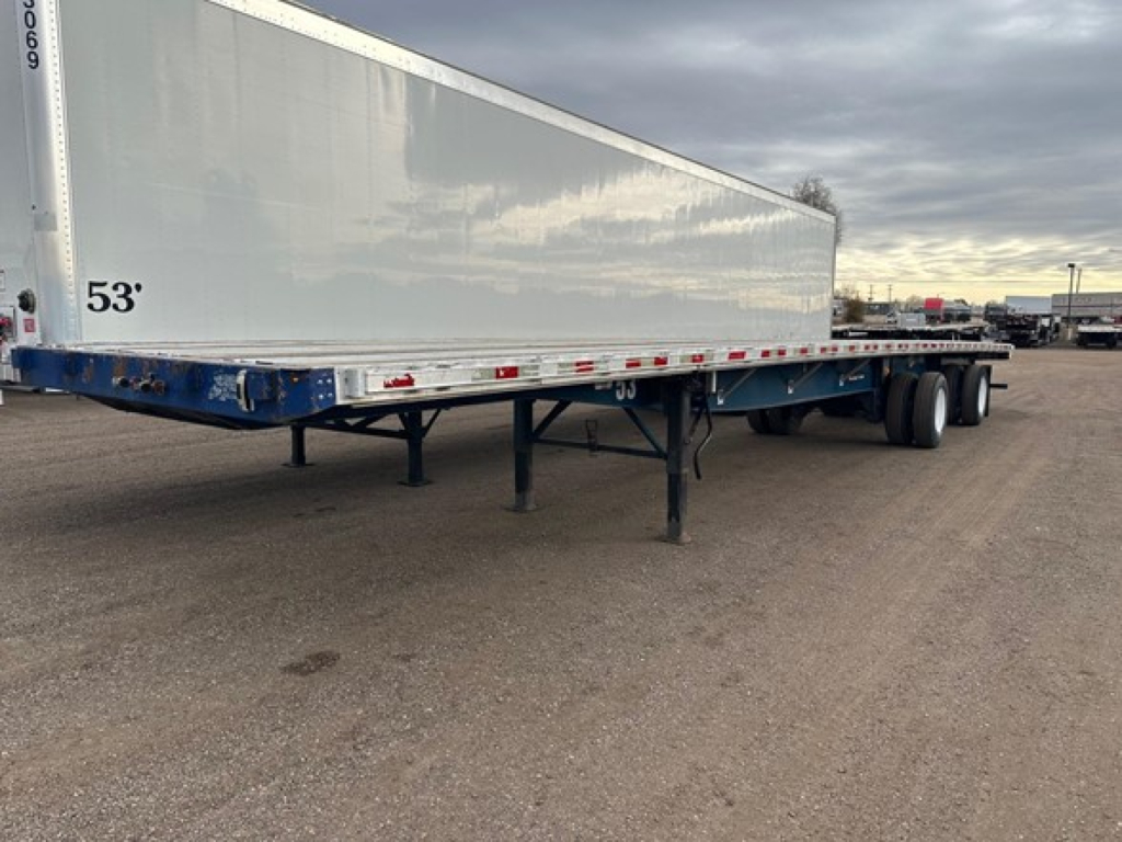 Flatbed trailer with blue undercarriage and white side panels, 53' length, dual rear axles, landing legs visible, storage area in a dealership lot.