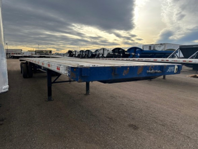 Flatbed trailer in a storage yard showing a blue frame with loading deck and tandem wheels undercarriage.