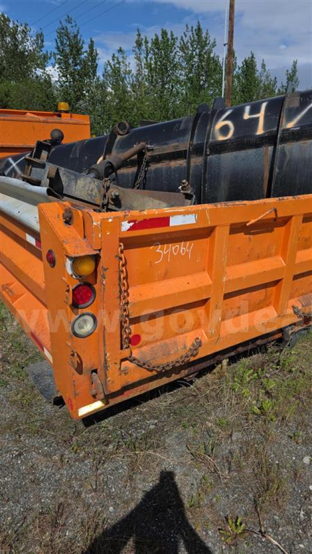 Back view of an orange Freightliner dump bed attached to a black container, with serial numbers and graffiti visible on the metal surfaces.
