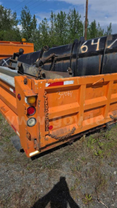 Back view of an orange Freightliner dump bed attached to a black container, with serial numbers and graffiti visible on the metal surfaces.