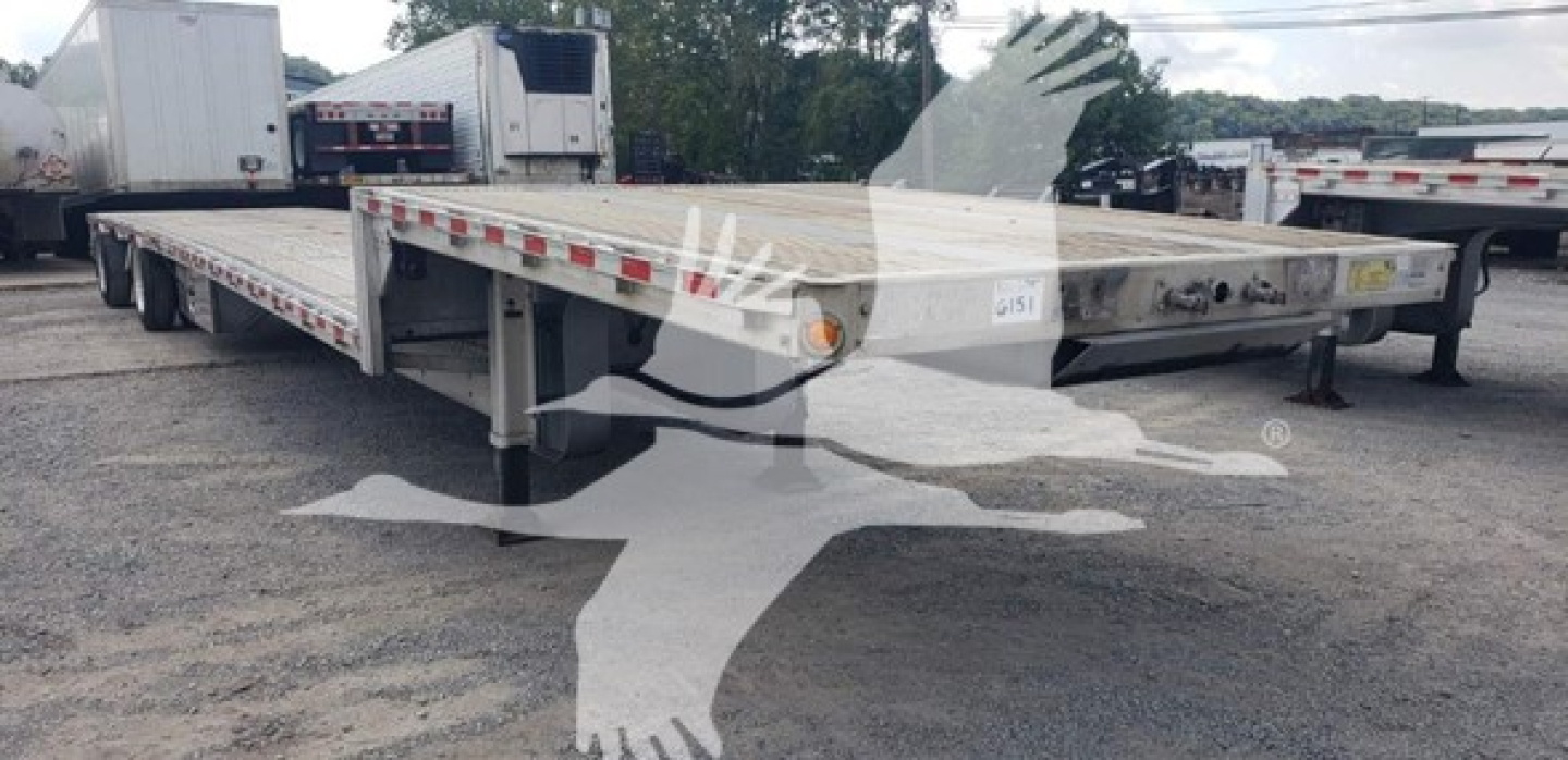 Flatbed trailer showing low deck with drop deck, metal surface, support legs, and reflector stripes along edge; visible wheels and adjacent trailers in a lot.