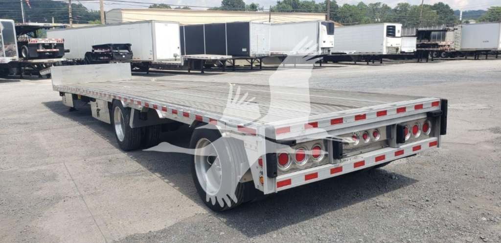 Flatbed trailer showing drop deck with dual rear axles and reflective tape along sides and rear; industrial yard in background.