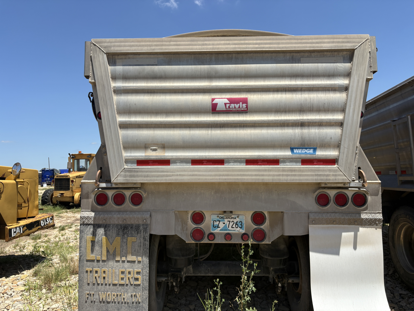 Dump trailer rear showing Travis logo on gray aluminum bed, taillights, bumper, and license plate.