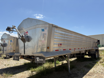 Dump trailer with metal side panels and front hitch, Travis logo visible, side railing, support legs down, American flag sticker, VIN label on rear.