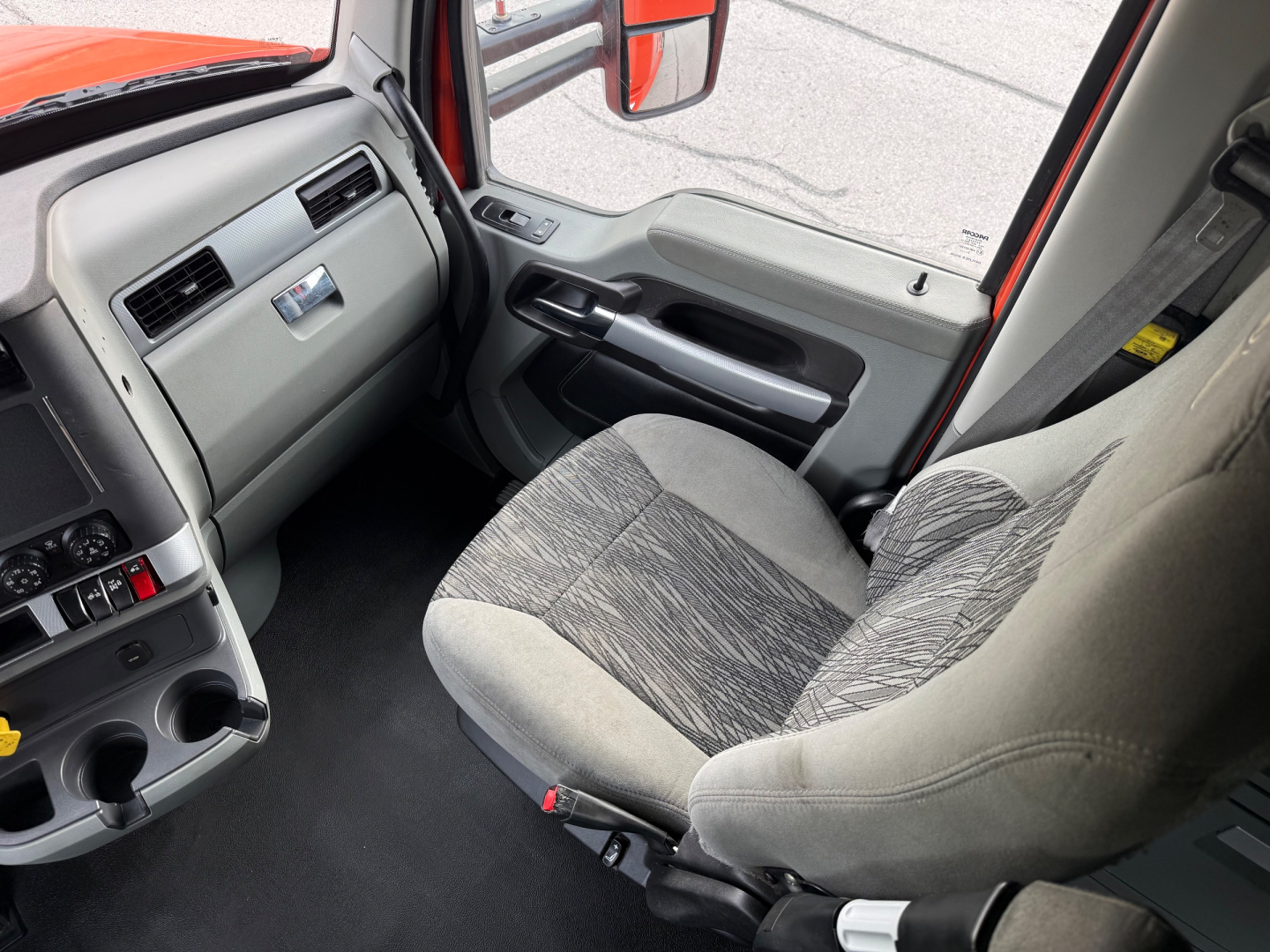 Cab interior of a 2021 Kenworth T680 showing driver seat, steering wheel area, dashboard with vents and storage compartments, grey fabric seat and door panel.