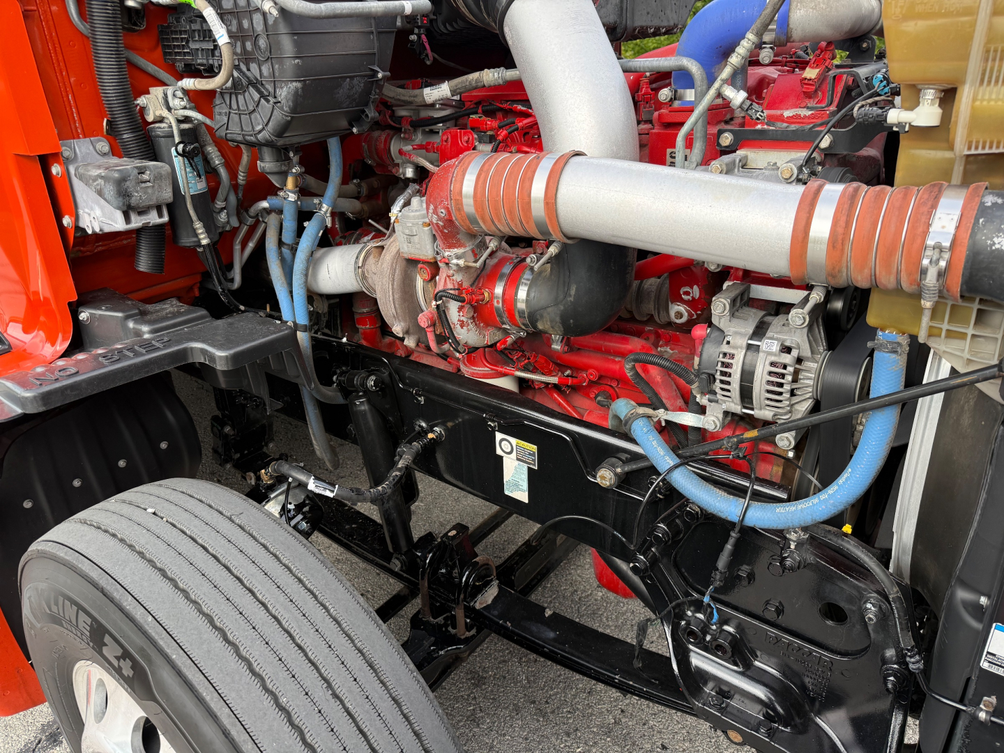 Kenworth T680 engine bay showing large turbo intake, red engine block, blue hoses, and black chassis with label placard.