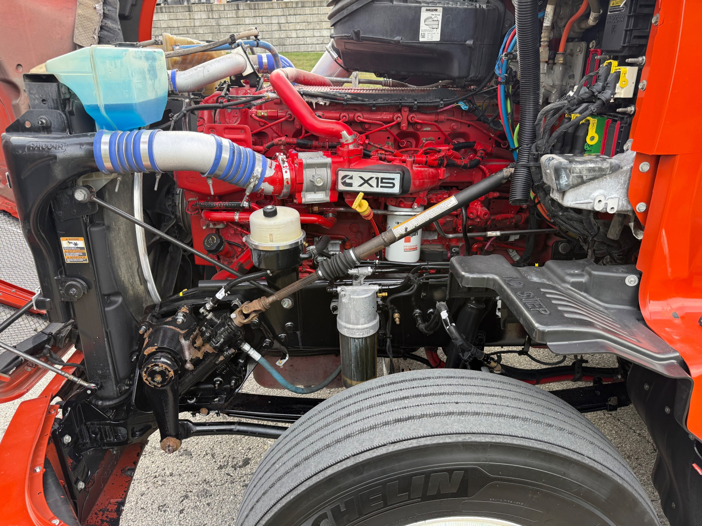 Kenworth T680 engine bay view with red 6.7L? engine labeled X15 turbo; visible coolant hoses, oil filter, and front suspension components.