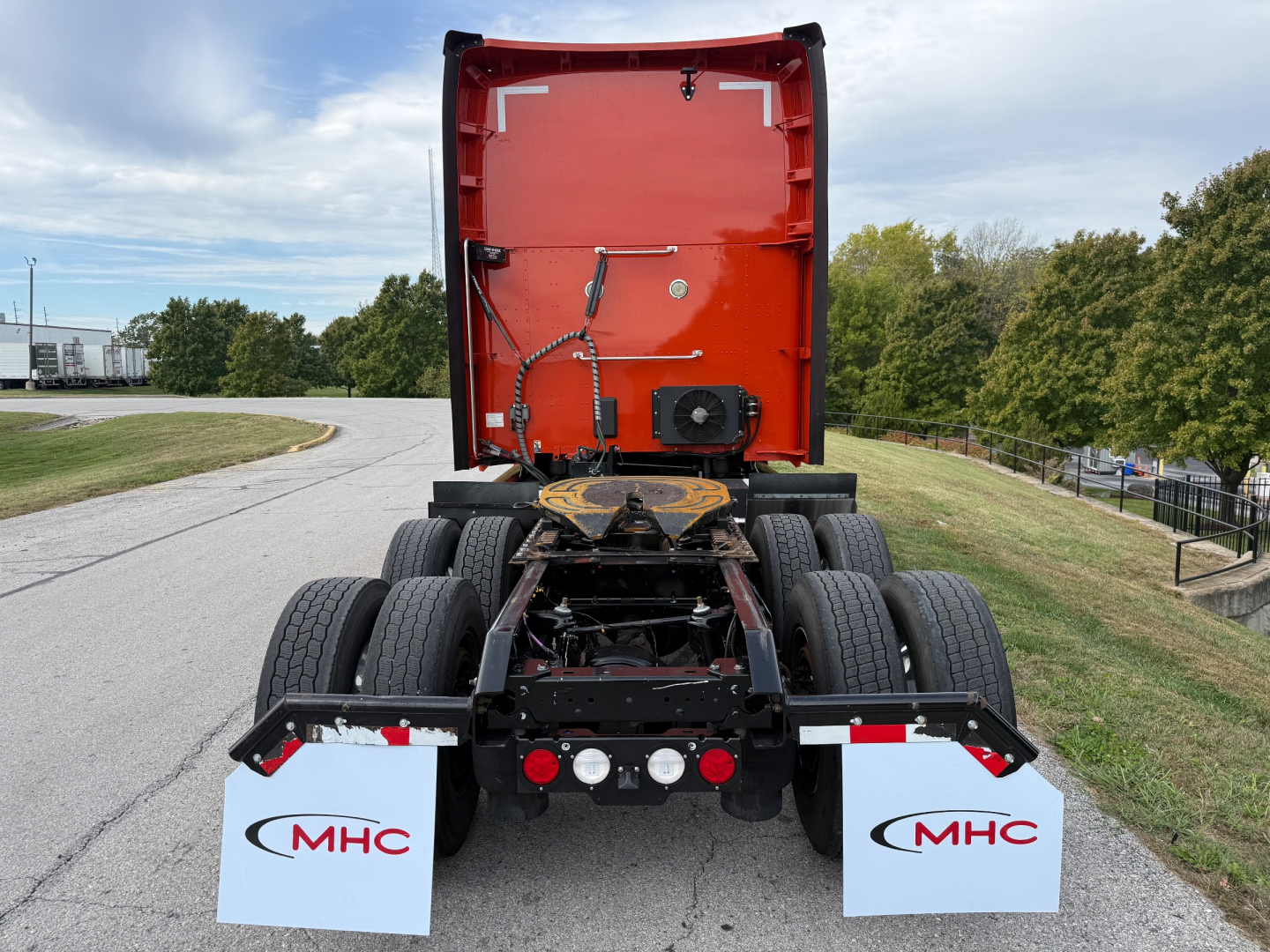 Kenworth T680 rear view showing chassis and fifth wheel area, orange cab back, twin rear axles, mud flaps with MHC logo, and tail lights.