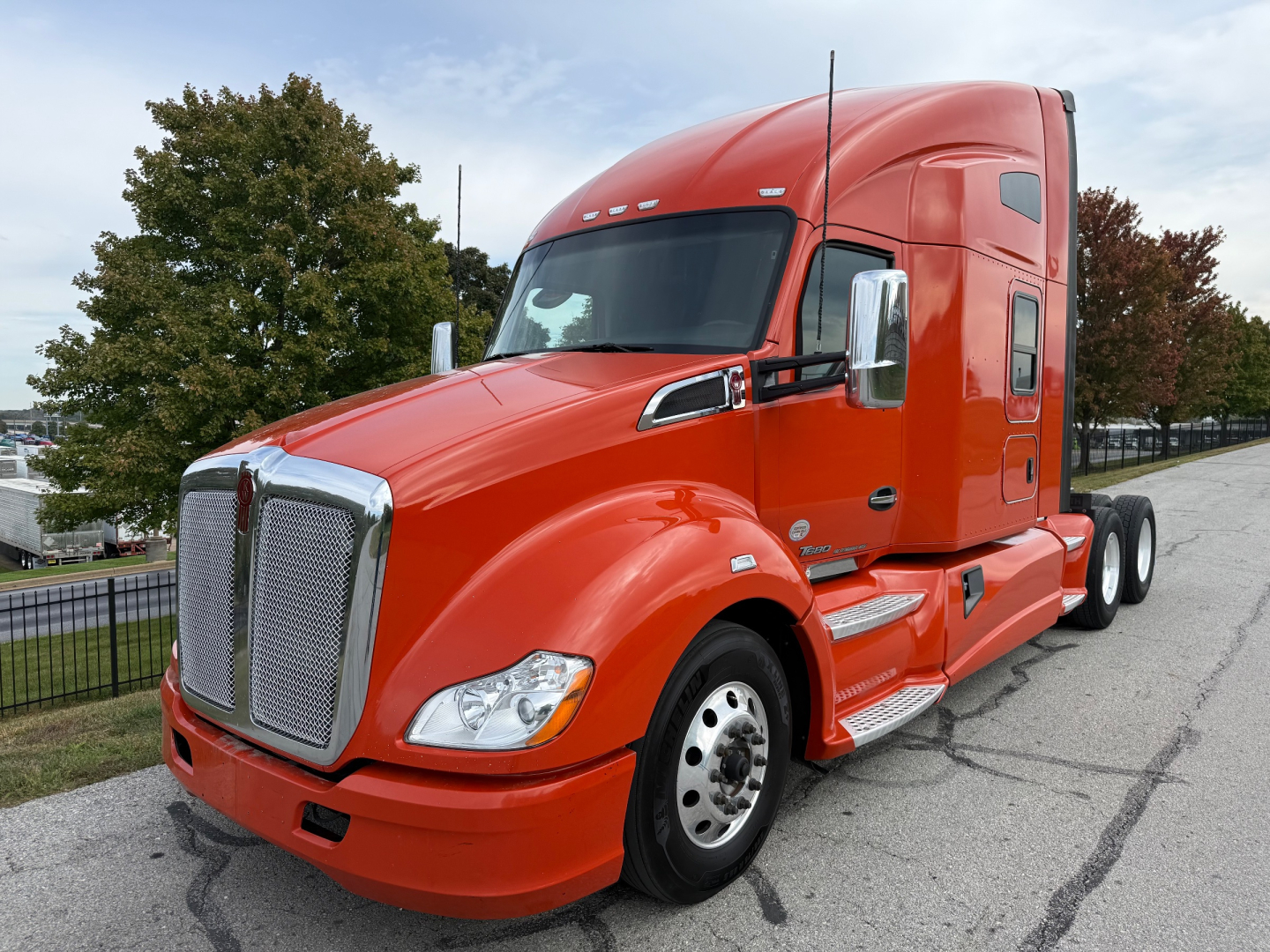 Kenworth T680 tractor cab in red viewed from front-left showing chrome grille and sleeper cab, with visible wheels and side steps.