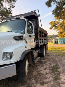 Truck cab and dump bed of a Freightliner 114SD with white cab and black dump body, front left angle, company name Warren on bed, dirt ground.