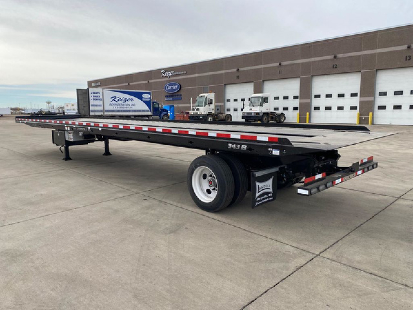 Container trailer flatbed showing Landoll branding and wheel area with single axle and dual tires, rear ramps and safety striping, with a building in background.