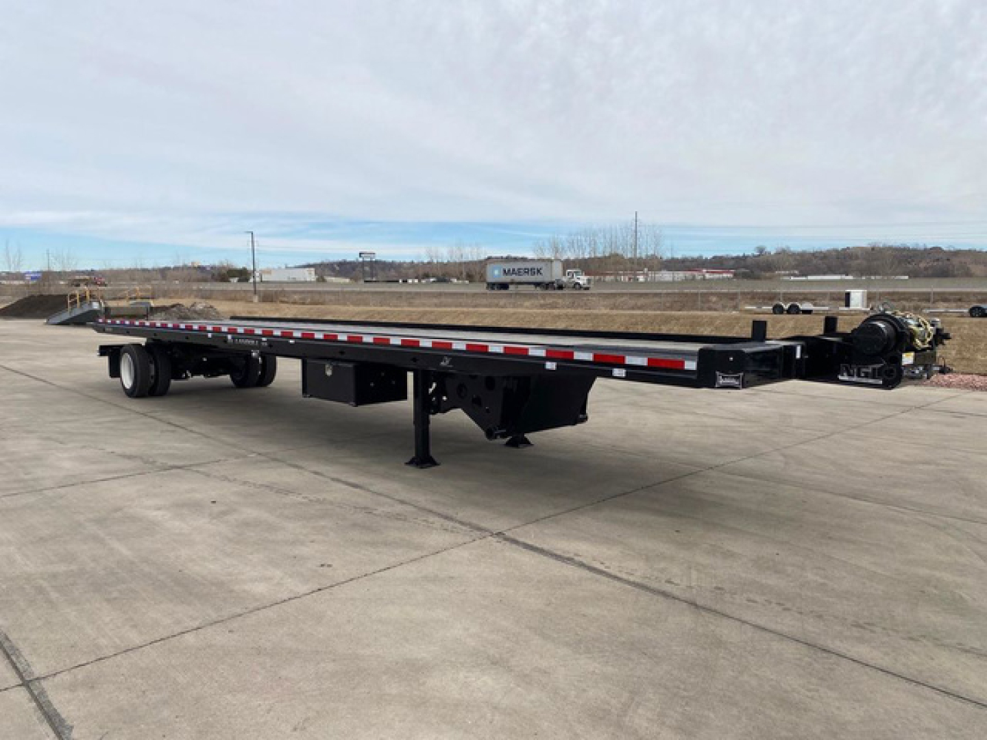 Flatbed trailer for container transport showing long black frame, tandem axles, landing gear, and gooseneck hitch.