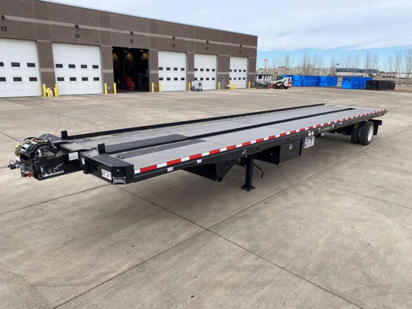 Flatbed trailer for trucking shows long steel deck with wheelset at rear, gooseneck hitch on left, safety rails and reflective striping along sides; outdoor yard with storage bays.