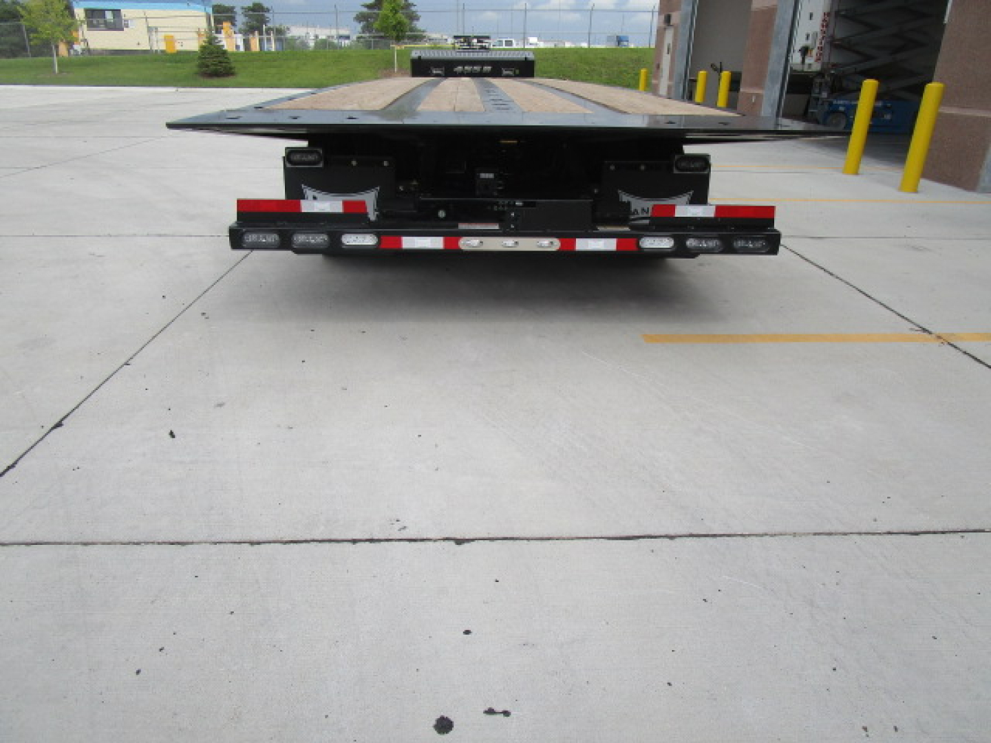 Flatbed trailer viewed from rear with Landoll 455 undercarriage, red and white reflective strips, loading platform extended slightly, industrial building backdrop.