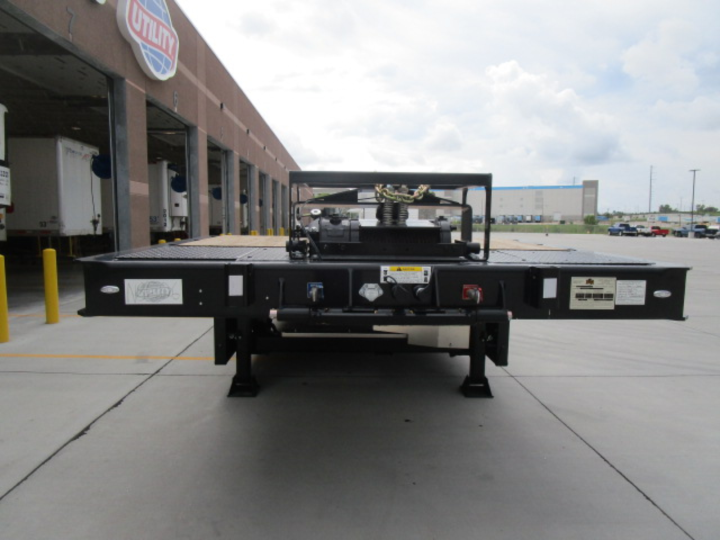 Flatbed trailer neck showing controls, hydraulics, and label stickers with platform and axles visible in distance.