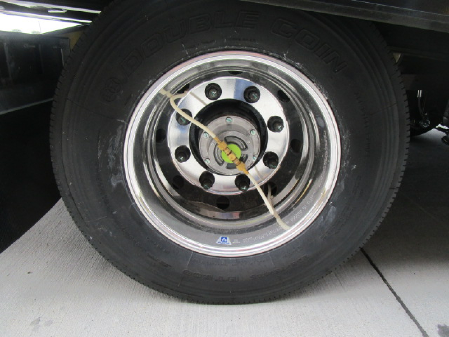 Wheel of a trailer showing a steel rim with multiple lug nuts and a green/yellow center hub; black tire with tread labeled ‘G2 CON’ visible; safety wire across wheel; blue and white certification label near rim.