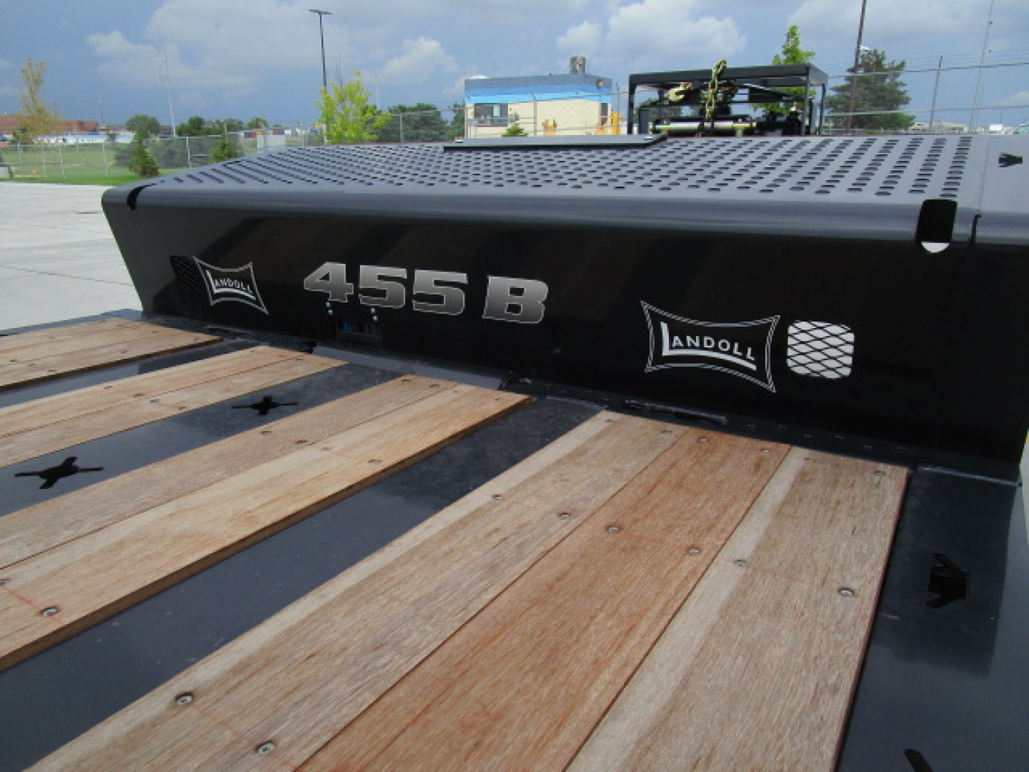 Flatbed trailer deck with black side panel labeled '455B' and 'Landoll' logos; wooden planks on bed and perforated metal upper surface visible.