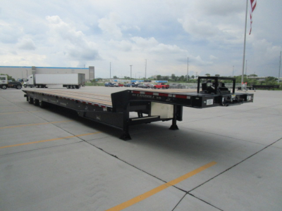 Flatbed trailer in an outdoor lot showing long black frame with mid-section ramps and gooseneck, no loading equipment visible.