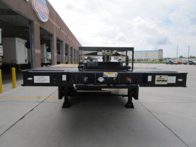 Flatbed trailer neck showing controls, hydraulics, and label stickers with platform and axles visible in distance.