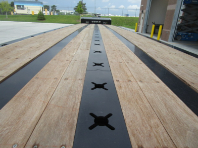 Long Landoll trailer deck with wooden planks and black center strips featuring cross-shaped tie-down anchors; distant building and equipment visible at trailer rear.