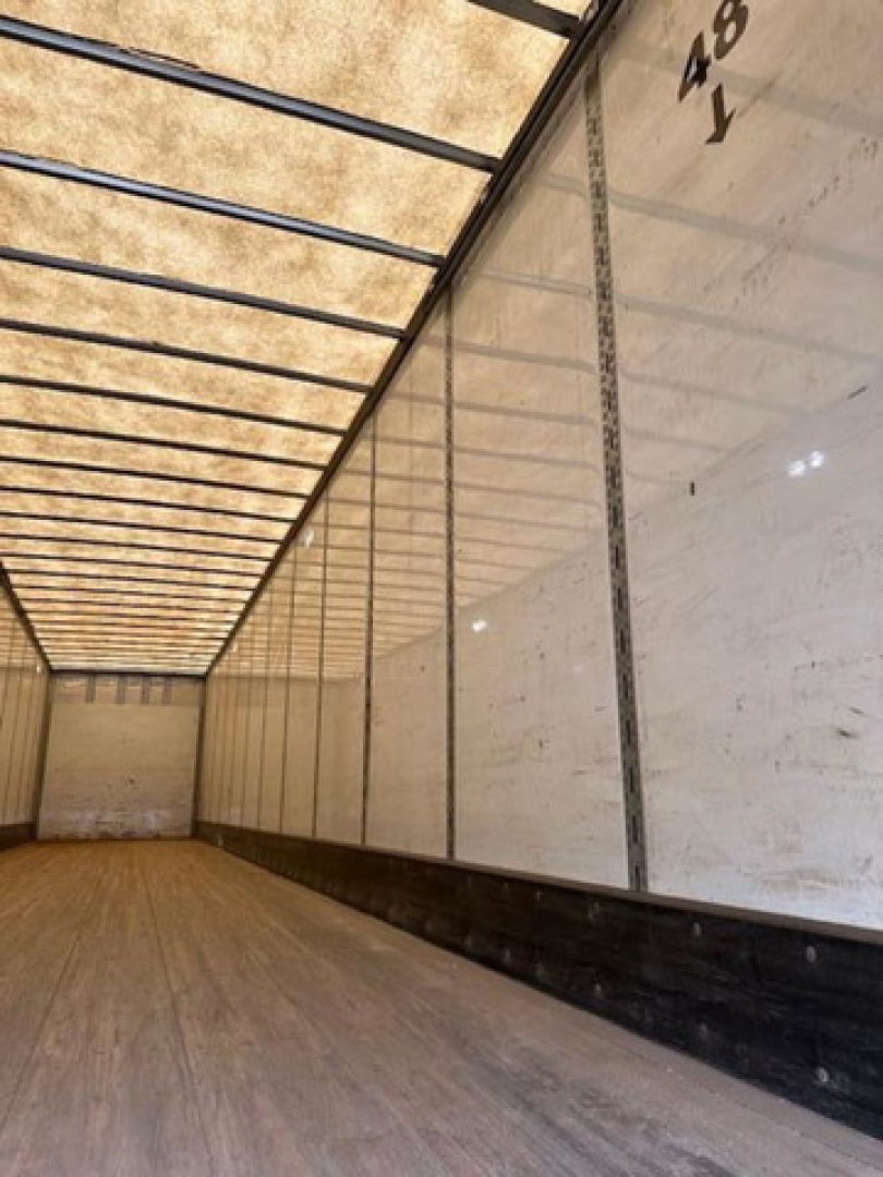 Interior of a cargo trailer with wooden floor and white side panels; exposed ceiling ribs; faint markings '48' and 'Y' on the wall.