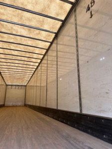 Interior of a cargo trailer with wooden floor and white side panels; exposed ceiling ribs; faint markings '48' and 'Y' on the wall.