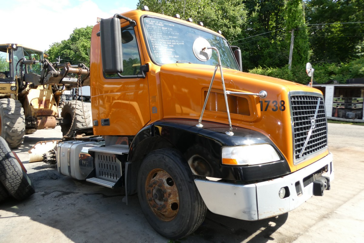 Orange Volvo VNM42T conventional daycab truck, front-right view, with large grille, black fenders, white bumper, and worn front left tire; VIN tag visible.